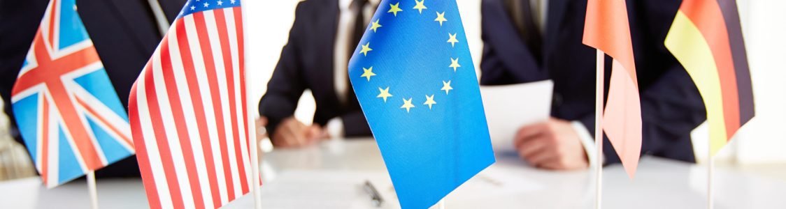 Image of several flags of different countries on workplace with three partners negotiating on background